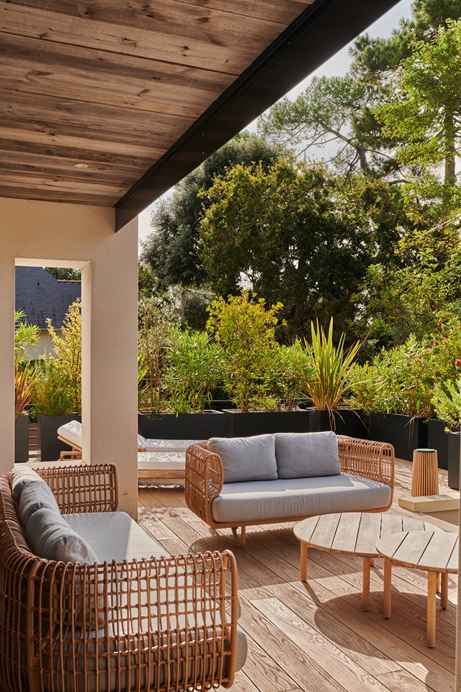 Terrasse ensoleillée, salon de jardin en osier et coussin en tissu gris. Végétation en arrière plan.