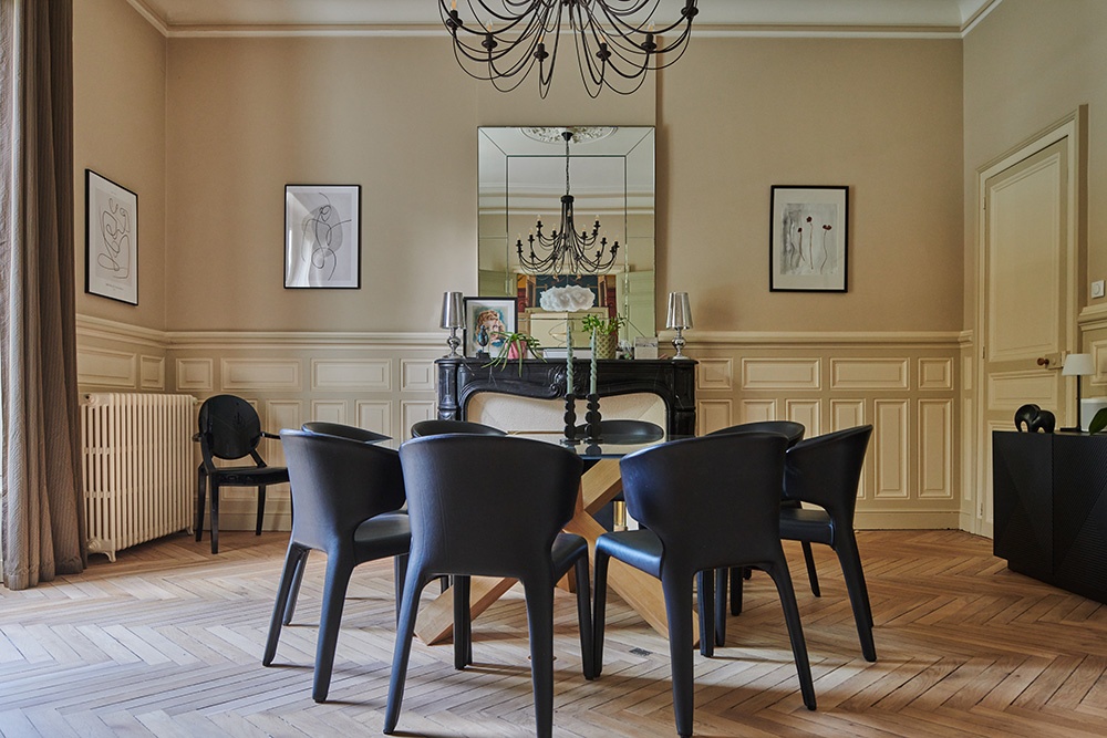 Salle à manger vintage, ancien parquet en chevron. Moulure sur les murs. Murs couleur crème et mobilier noir. Cheminée en marbre noir derrière la table à manger.