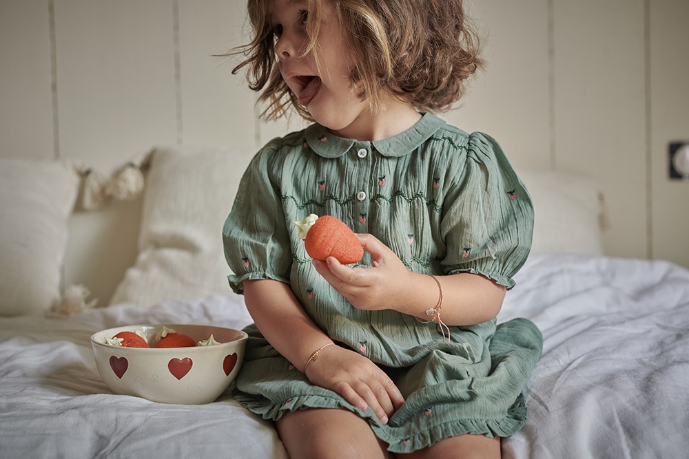 Fillette de 3/4 ans portant une robe verte brodé de petites fraises, un petit bracelet précieux, tient un bonbon en forme de fraises. Elle se tient assise sur un lit et regarde hors champ.