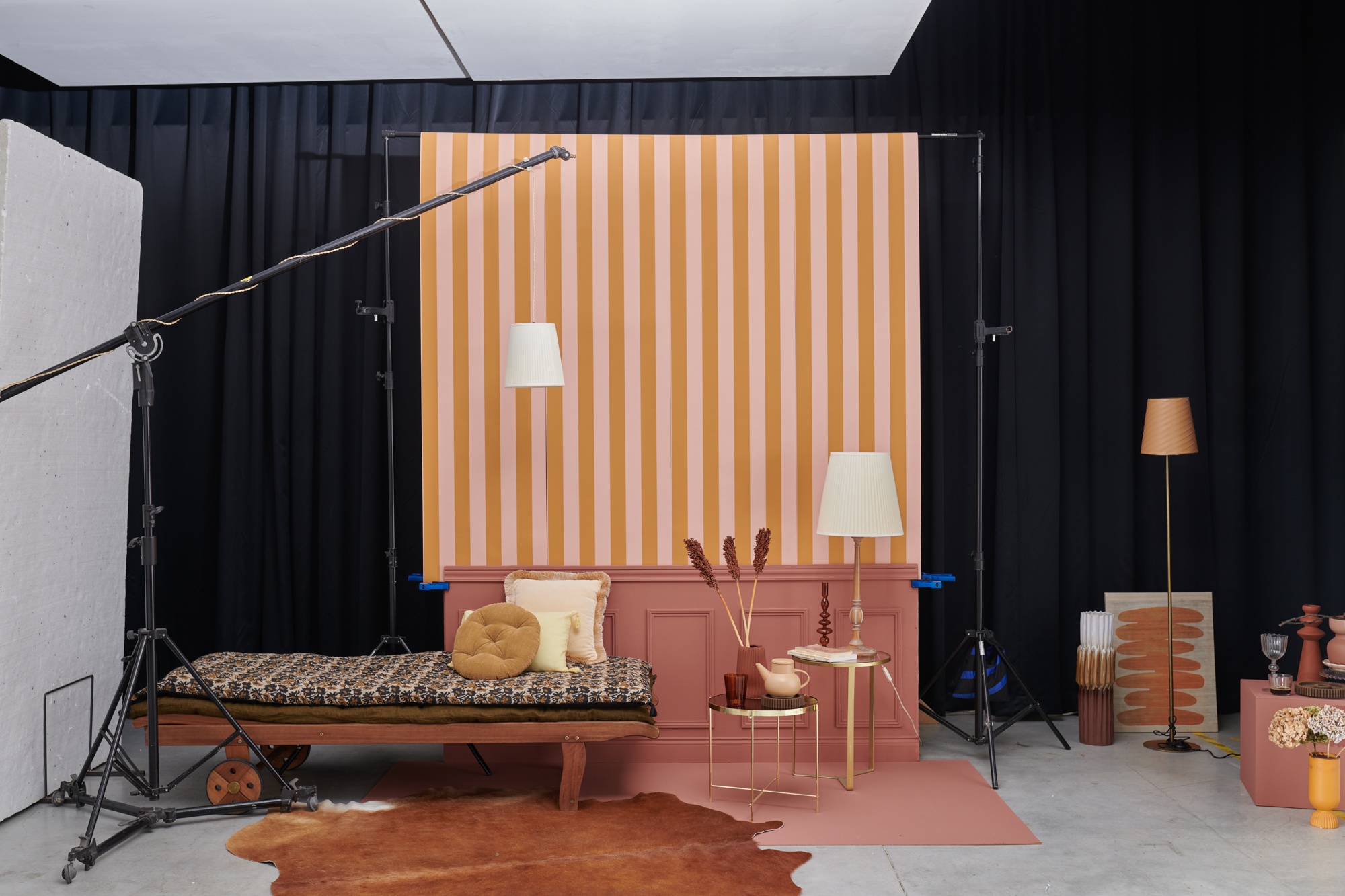 Décors dans un espace cyclo d'un studio de photographie. Du matériel photo un set design composé d'un fond principal rose à rayures oranges, soubassement de moulures rouges et d'accessoires de décoration : méridienne, luminaire suspendu, table de chevet circulaire or