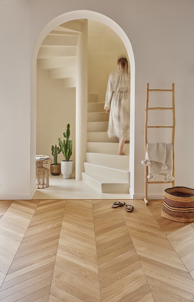 Une image lifestyle : une arche blanche intérieure donne à voir une femme de dos montant un escalier en colimaçon. Au 1er plan , le parquet en bois PANAGET vient donner une atmosphère douce.