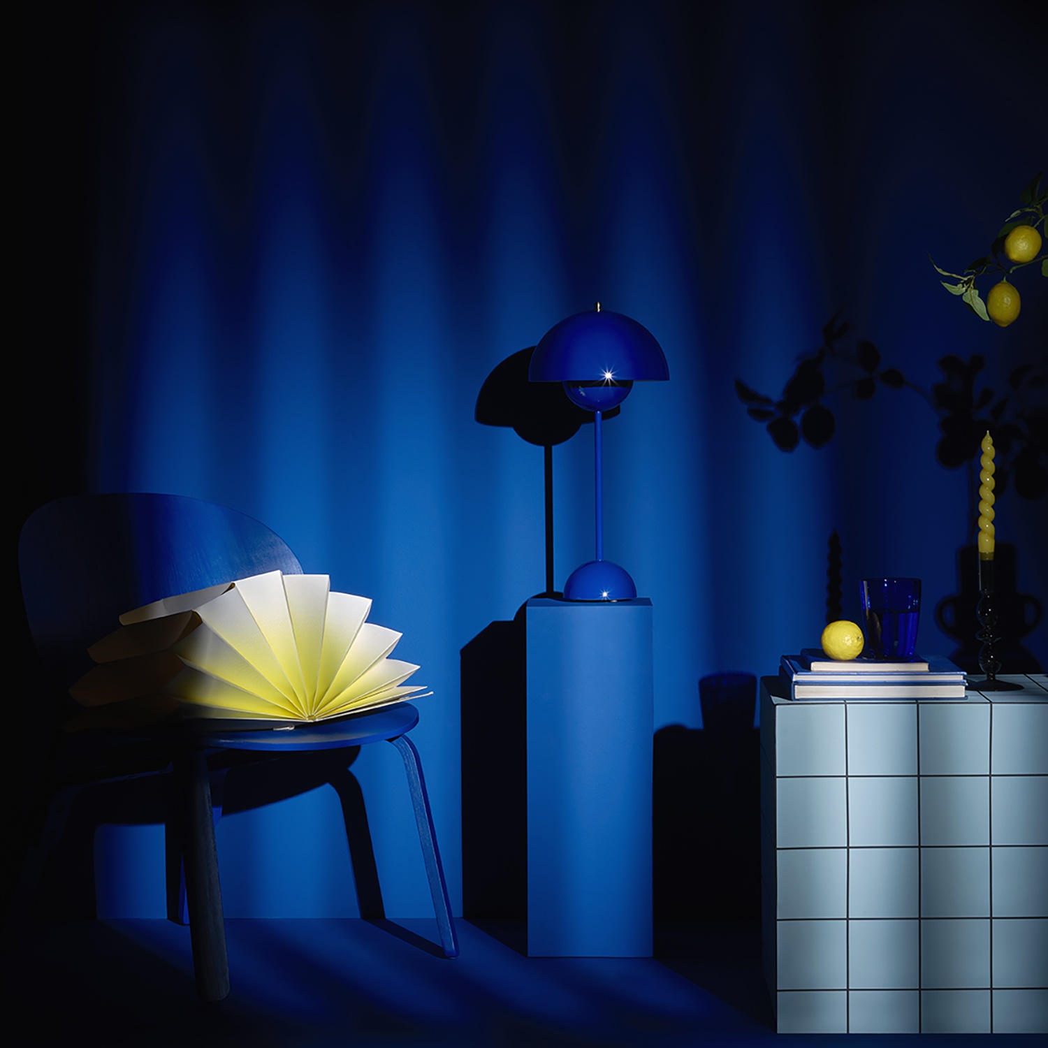 une nature morte comprenant une chaise, une table bleue, d'autres objets décoratifs très graphiques bleus cobalt et jaunes pastels/ La lumière de l'image joue le clair-obscur