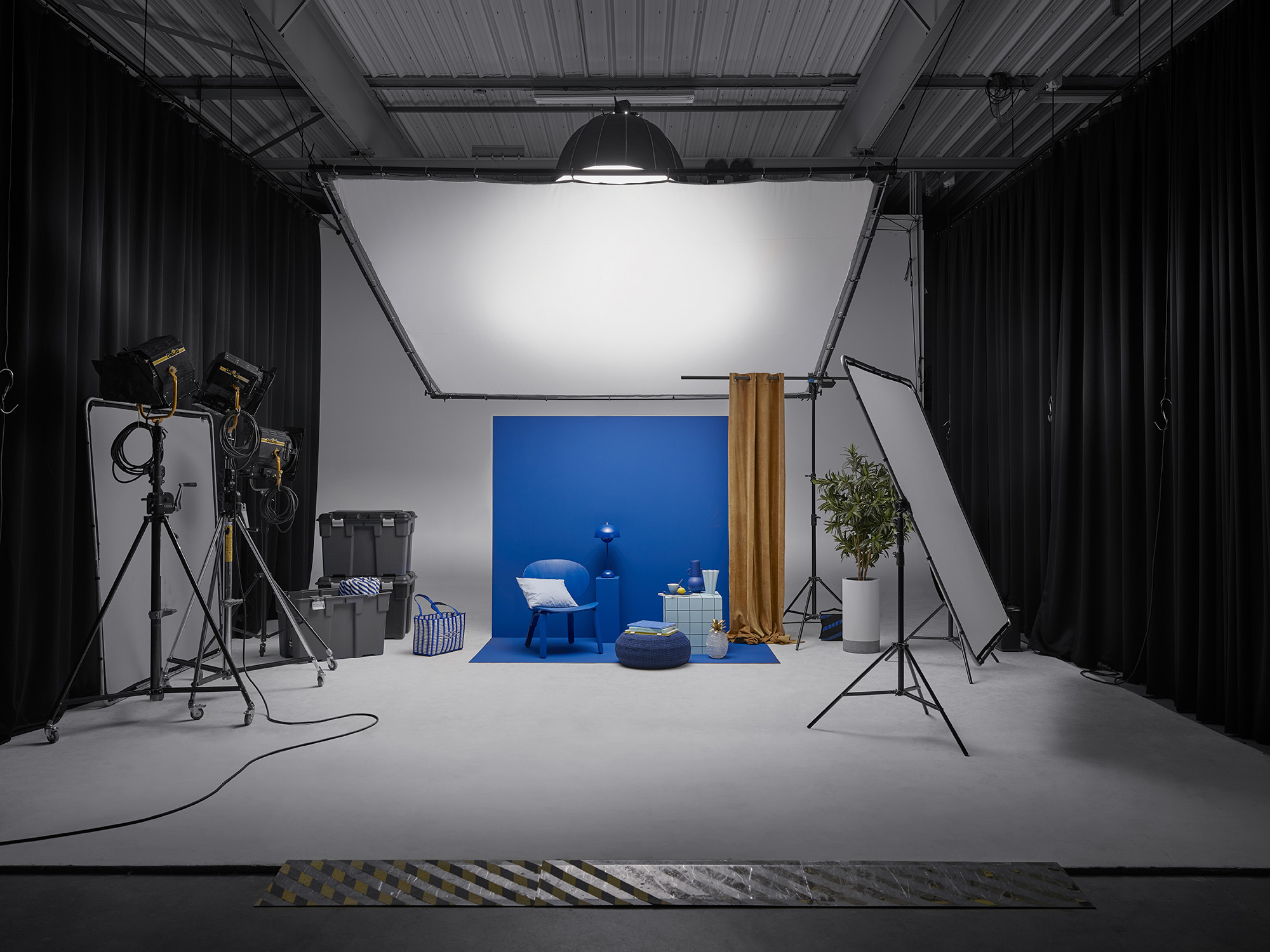 Décors dans un espace cyclo d'un studio de photographie. Du matériel photo un set design composé d'un fond principal lilas et d'accessoires de décoration : lampe bleue, rideau ocre, chaise colorée ...