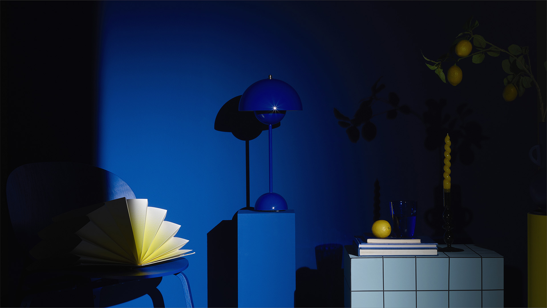une nature morte comprenant une chaise, une table bleue, d'autres objets décoratifs très graphiques bleus cobalt et jaunes pastels/ La lumière de l'image joue le clair-obscur