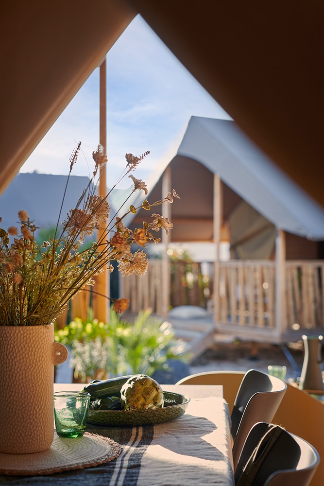 Plan serré depuis un intérieur de tente lodge de la marque SUNSHINE, un bouquet de fleurs fraiches posé sur la table : l'espace s'ouvre sur l’extérieur. L'ambiance est lumineuse.