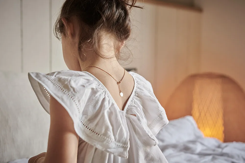 Fillette photographiée de dos, cadrage serré. La fillette porte une robe romantique de coton blanc et un délicat collier de la marque Bijoux Sucre d'Or